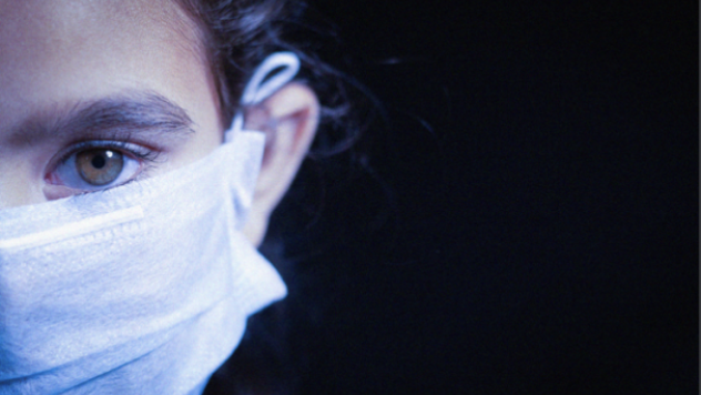 A person wearing a medical mask stares directly into the camera at close range with a black background.