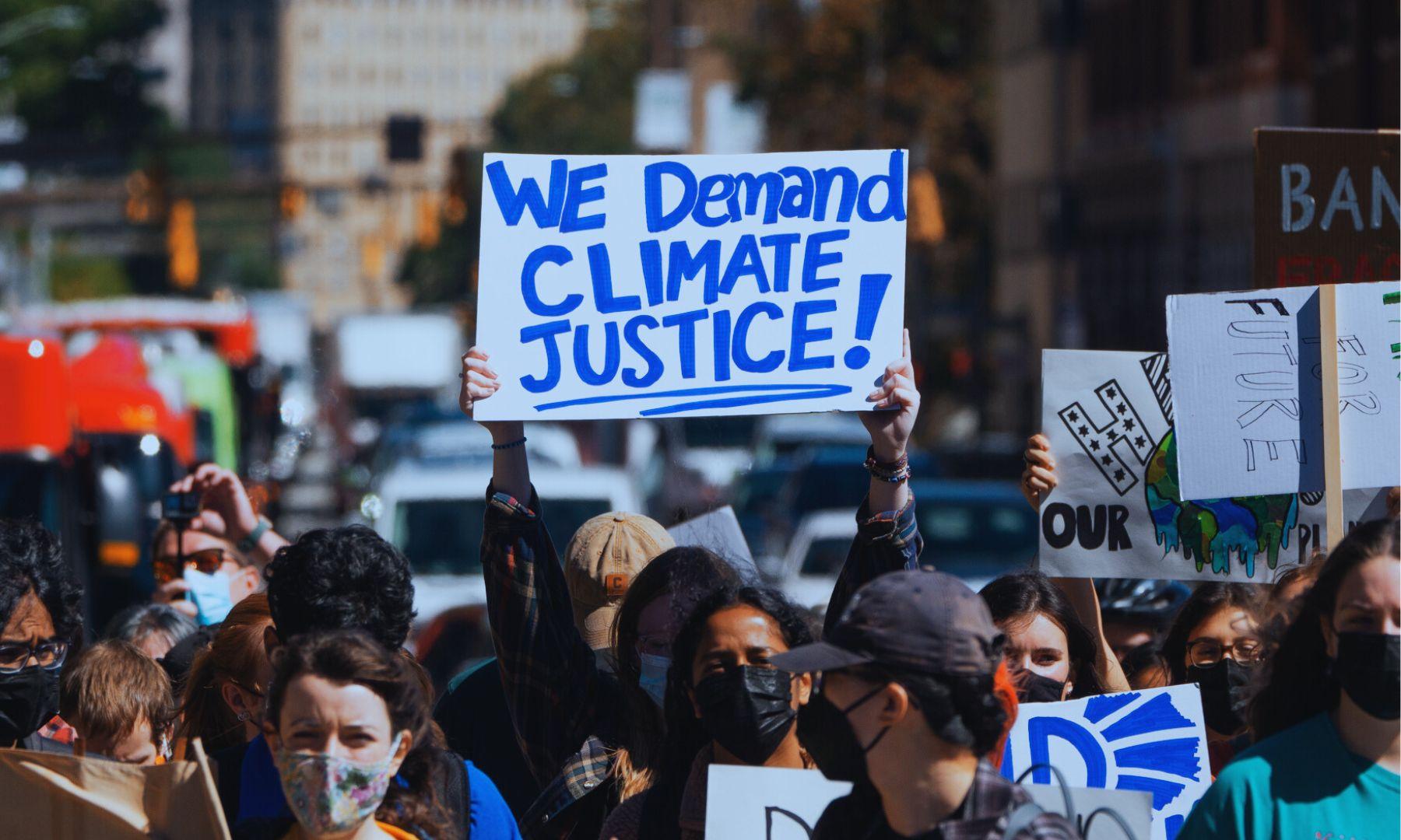 Person holding up sign in a group of people. Sign reads we demand climate justice now in blue letters on a white bristol board.