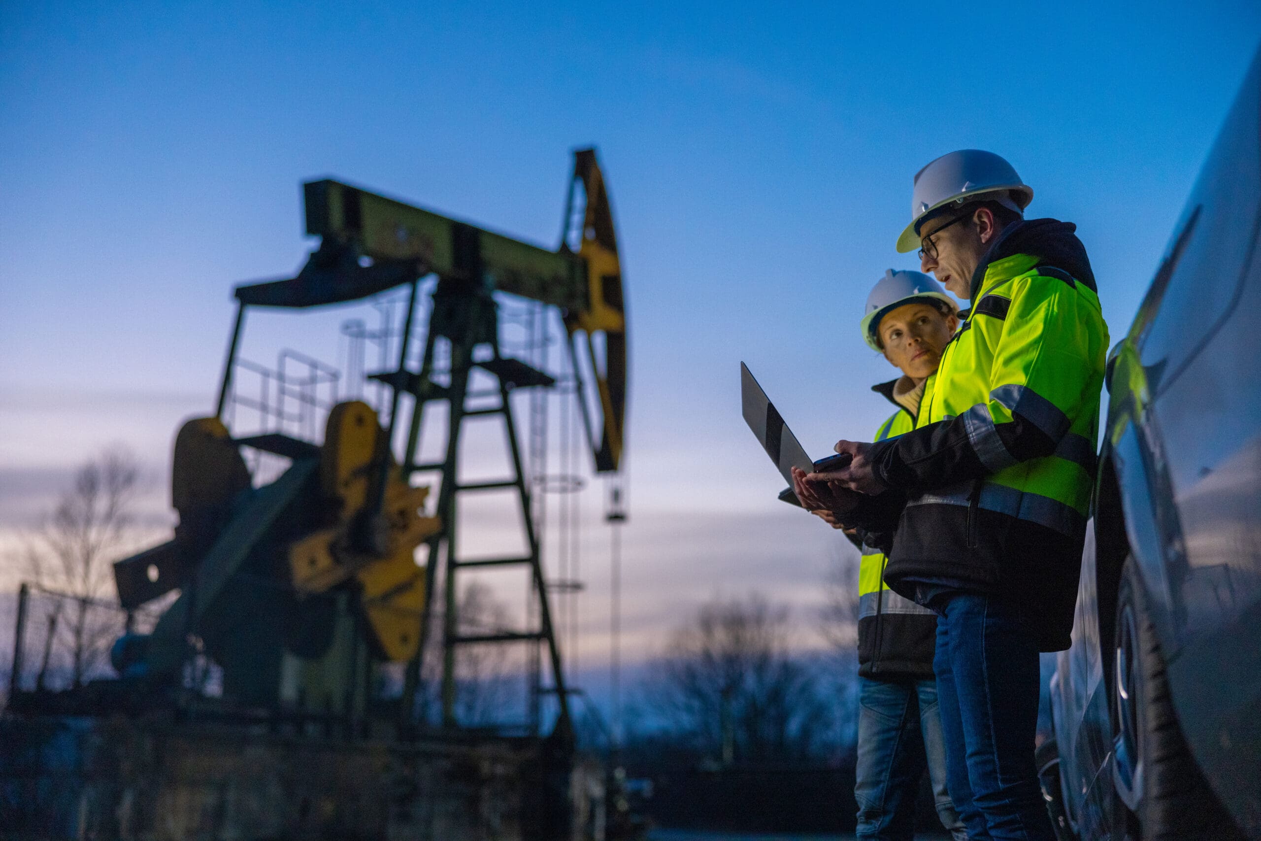 Low angle view of male and female partners in protective clothing planning over laptop while standing against equipment by car at field against sky during sunset