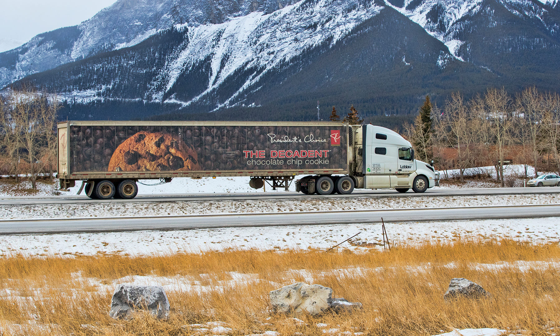 Transport truck on the highway with mountains in the background. Truck features an image of a chocolate chip cookie with chocolate chips in the background with the text President's Choice.