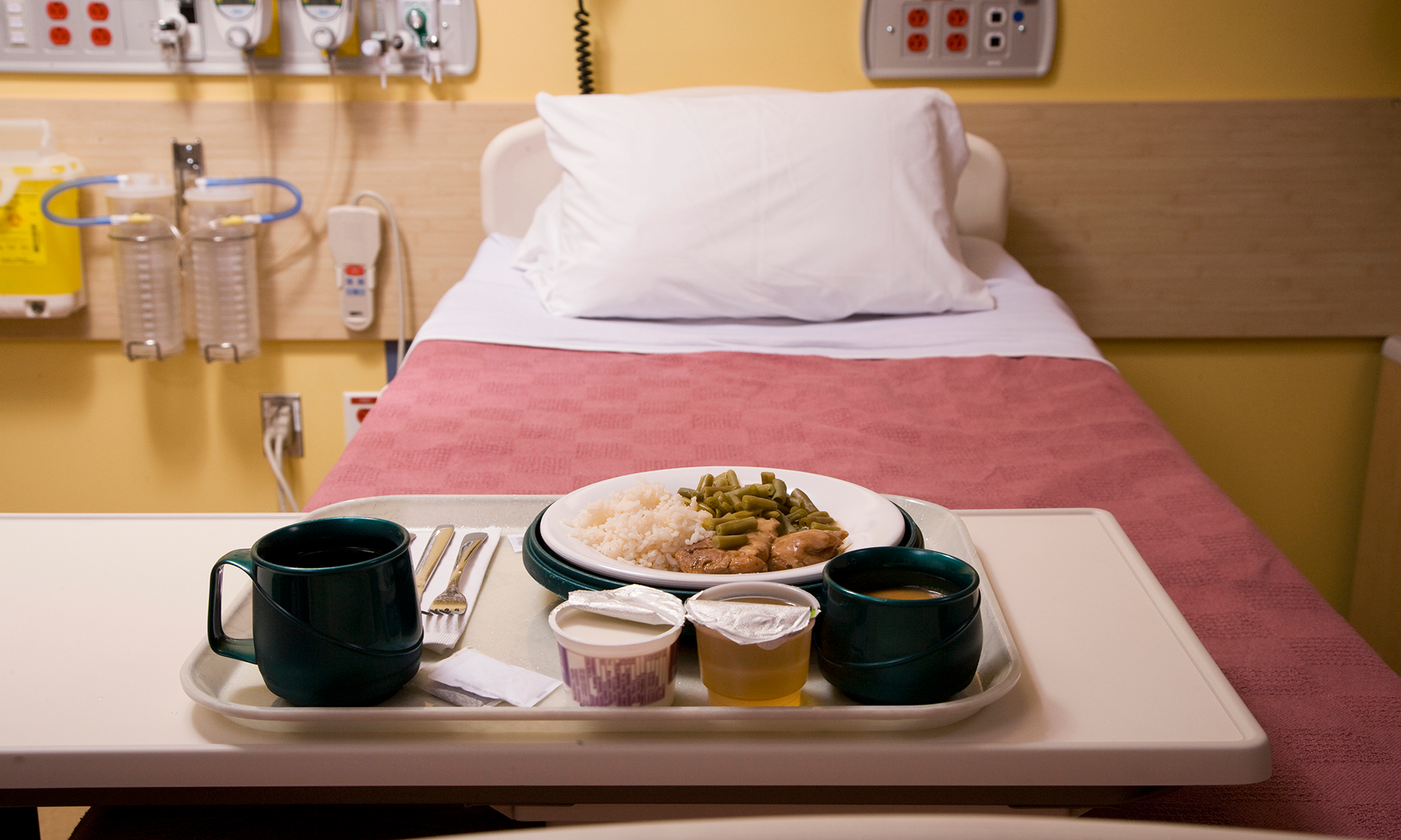 Empty hospital bed with pink blanket on a made bed. Food tray at the bottom of the bed with plate of food and drink